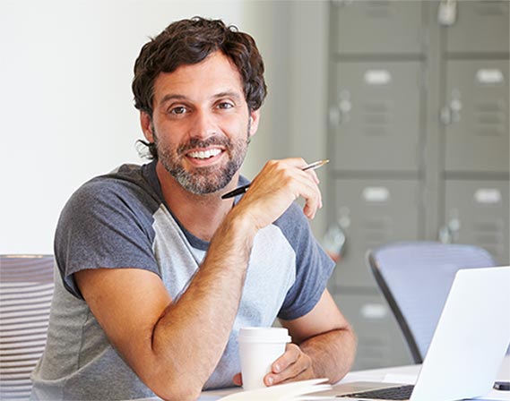 A man seated on front of laptop holding a pen in one hand and cup in another hand.